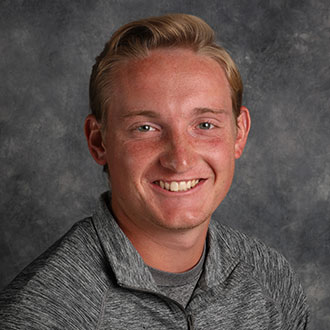 Michael Janes, Head Tennis Coach for the Athletics Department, stands in front of a mottled gray background.