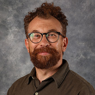 Jason Jaspersen, Associate Professor for the Studio Art Department, smiles in front of a gray textured background.