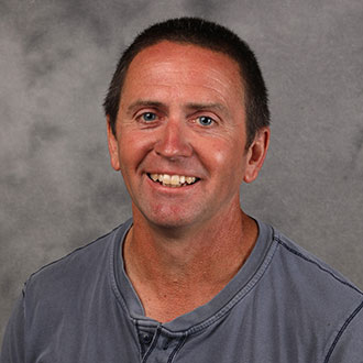 Dan Johnson, Head Groundskeeper for the Groundskeeping Department, smiles while posing in front of a gray studio backdrop.