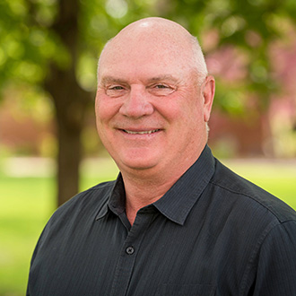 Ken Mueller, Head Clay Target Coach for the Athletics Department, stands outdoors on the Bethany Lutheran College campus, with green trees in the background.