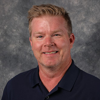 Christopher Kind, Director of the Advancement Department, is posed in front of a gray textured background.