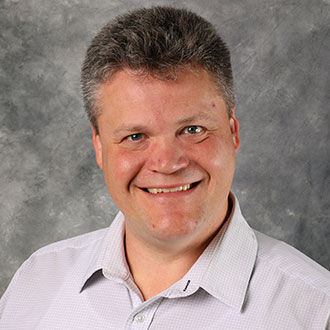 Brian Klebig, Professor for the Communication Department, stands in front of a gray marbled background.