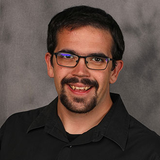 Zay Koepsell, Assistant Professor and Instructor for the Business Administration Department, smiling in front of a gray background.