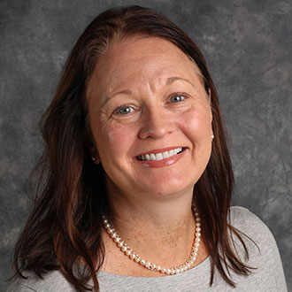 Jen Kom, Professor in the Psychology Department, smiles in front of a gray mottled background.