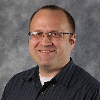Jon Kovaciny, Web Development Specialist for the Institutional Communication Department, stands in front of a gray mottled background.