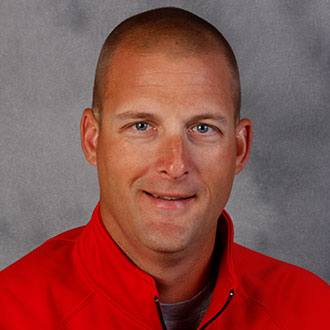 Ryan Kragh, Co-Athletic Director and Head Baseball Coach for the Athletics Department, smiling in front of a gray background.