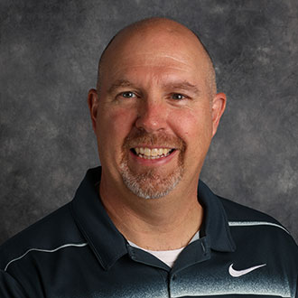 Andy Krueger, Associate Professor for the Business Administration Department, poses in front of a gray mottled background.