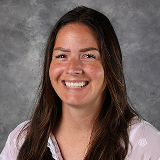 Alanna Lienig, Associate Professor for the Education Department, smiles in front of a gray mottled background.