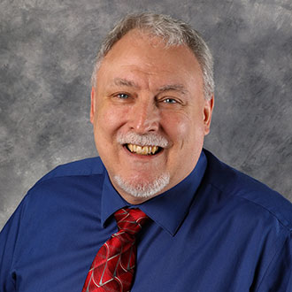 Perry Lund, Assistant Professor for the Computer Science Department and Adjunct Faculty for the Physics Department, posed against a gray mottled background.