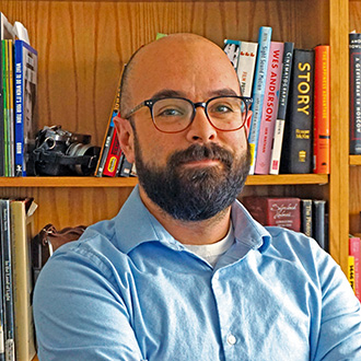 Ben Lundsten, Assistant Professor, Media Arts Department with glasses and a beard, wearing a blue shirt, stands in front of a bookshelf filled with books and a camera.