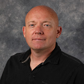 Derick Lyngholm, Associate Professor for the Communication Department and Head Coach Men's Soccer for the Athletics Department, posing in front of a gray mottled background.