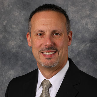 Ted Manthe, Vice President of the Student Affairs Department, Title IX Coordinator for the President's Office Department, and Instructor of Teaching Christian Faith for the Education Department, poses in front of a gray textured background.