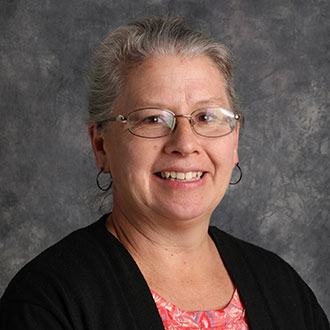 Laura Matzke, Instructor of Organ, Piano, and Voice for the Music Department, standing in front of a gray backdrop.