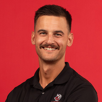 Mitch DeKruif, Head Golf Coach with short dark hair and a mustache, smiling while wearing a black collared shirt, standing against a red background.