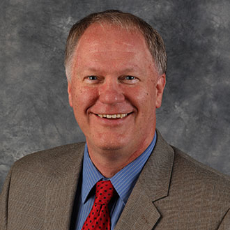 Daniel Mundahl, Senior Vice President of Finance and Administration of the Finance and Administration Department and Online Instructor for the Broad Field Social Studies Department, stands in front of a gray textured background.