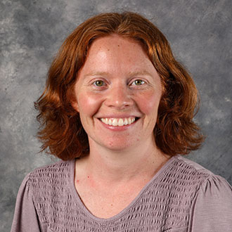Allison Petzel, Online Instructor of the Biology Department, smiles in front of a gray mottled background.