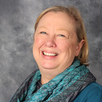 Carrie Pfeifer, Professor, Education Department, wearing a teal scarf and blue top, posing in front of a gray backdrop.