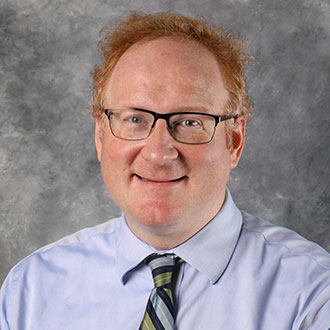 Nick Proksch, Adjunct Faculty for the Languages Department, stands in front of a gray background.