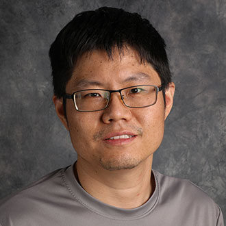 Sheng Sang, Assistant Professor of the Engineering Sciences Department, softly smiles in front of a gray textured background.