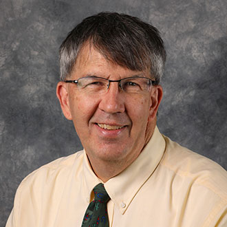 John Sehloff, Director of the Information Technology Services Department and Online Instructor in the Computer Science Department, smiling in front of a gray backdrop.