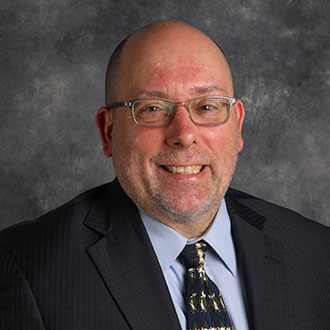 Jonathan Stadler, Professor of the Psychology Department, smiles while posed in front of a gray textured background.