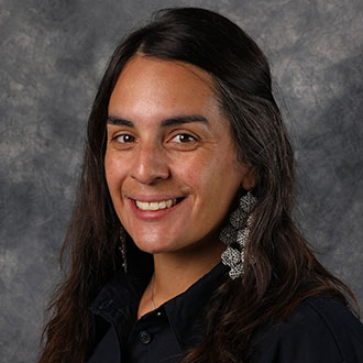 Helena Stevens, Associate Professor of the Clinical Mental Health Counseling, Graduate Programs Department, smiling in front of a gray mottled background.