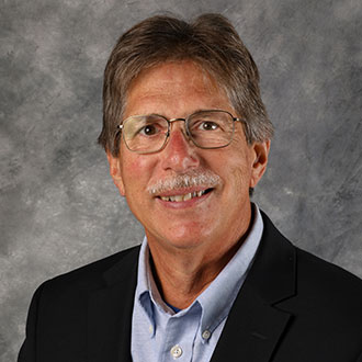 Dave Urness, String Instructor of the Music Department, smiles in front of a gray textured background.
