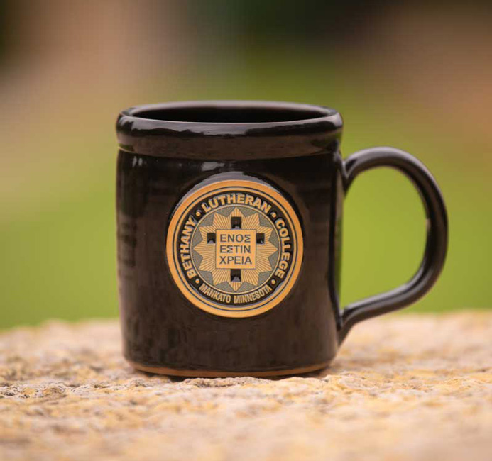 A black mug featuring the Bethany Lutheran College seal rests on a stone surface in Mankato, set against a blurred green background.