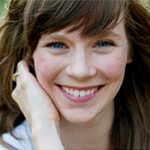 A Bethany Lutheran College student with long brown hair smiles at the camera, resting her hand on her cheek while sitting outdoors in natural light.