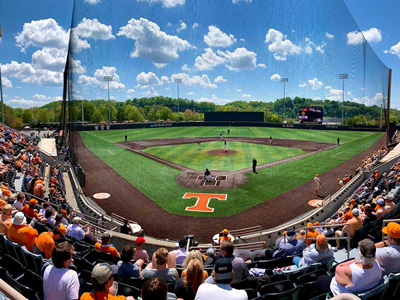 Bethany Lutheran College students watch a baseball game at the stadium with a large orange T behind home plate on a sunny day.