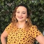 A Bethany Lutheran College student stands outdoors in front of green foliage, wearing a yellow patterned shirt and smiling at the camera.