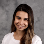A young woman with long brown hair smiles, wearing a white top, in front of a gray mottled background at Bethany Lutheran College in Mankato, Minnesota.