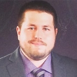This image features a man with short dark hair and a beard, dressed in a suit and tie, posed before a dark background in Mankato, Minnesota. If connected to Bethany Lutheran College, this could be a faculty or staff profile photo.