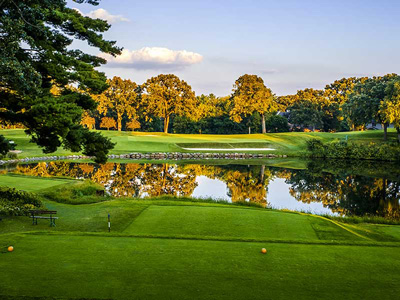 A scenic green golf course in Minnesota features a tranquil pond in the foreground, bordered by lush trees and set beneath a clear blue sky.