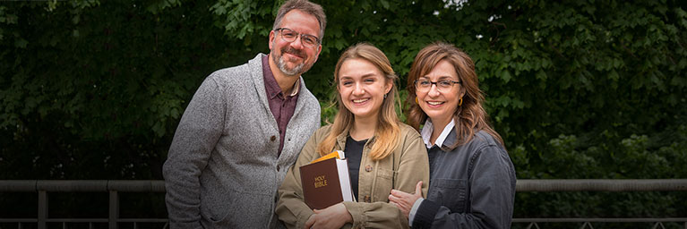 Three adults stand outdoors in Mankato, smiling for the camera. The person in the center holds a book titled Holy Bible. This scene may be related to a Bethany Lutheran College event or gathering.