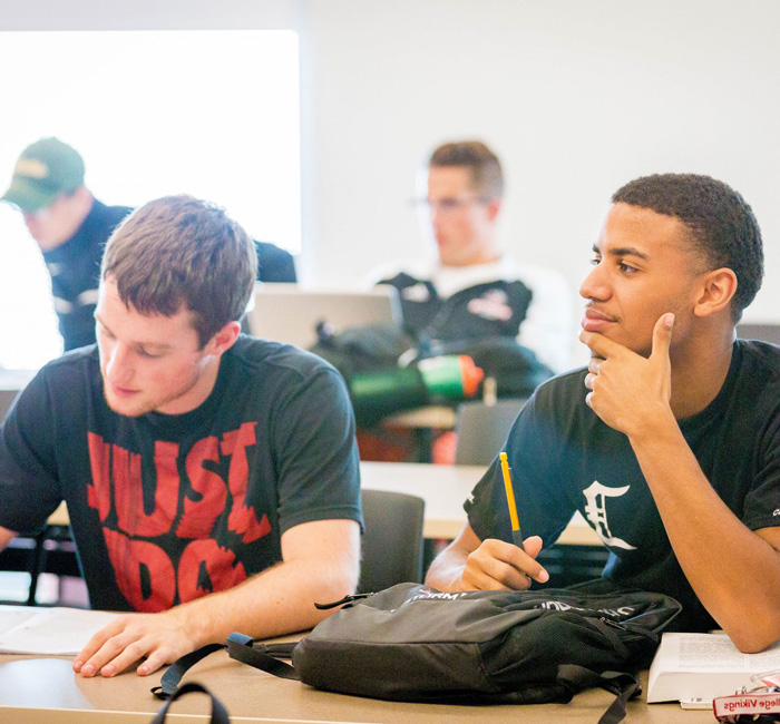Two Bethany Lutheran College students sit at desks in a Mankato classroom; one student writes while the other looks forward, holding a pencil.