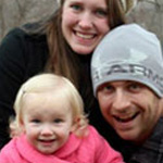 Three people smile at the camera outdoors at Bethany Lutheran College, Mankato: two students in hats stand behind a younger student in a pink coat.