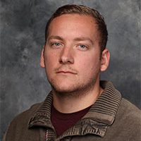 A Bethany Lutheran College student with short hair in a brown sweater poses against a gray, textured background.