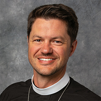 Kyle Madson, Adjunct Faculty wearing a clerical collar, with short brown hair, smiling at the camera against a gray mottled background.