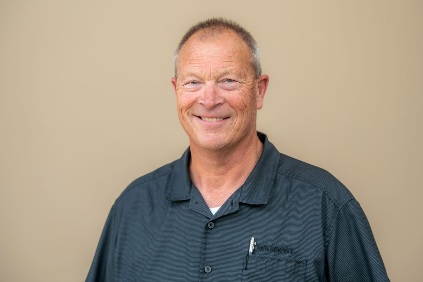A smiling older man with short gray hair wearing a dark gray collared shirt. He is standing in front of a plain beige background, looking directly at the camera. He is Joel Meyer, recipient of the 2025 Distinguished Alumni Award.