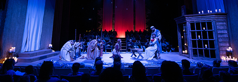 Stage production of a Nativity scene, lit in blue and red tones, with actors dressed in biblical costumes portraying Mary, Joseph, shepherds, and the baby Jesus. The audience surrounds the stage, which is decorated with candles, draped fabric, and rustic wooden structures.