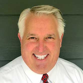 Man in a white shirt and red tie smiling against a dark paneled background.