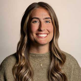 Portrait of a smiling woman with long, wavy brown hair in a tan sweater against a neutral background.
