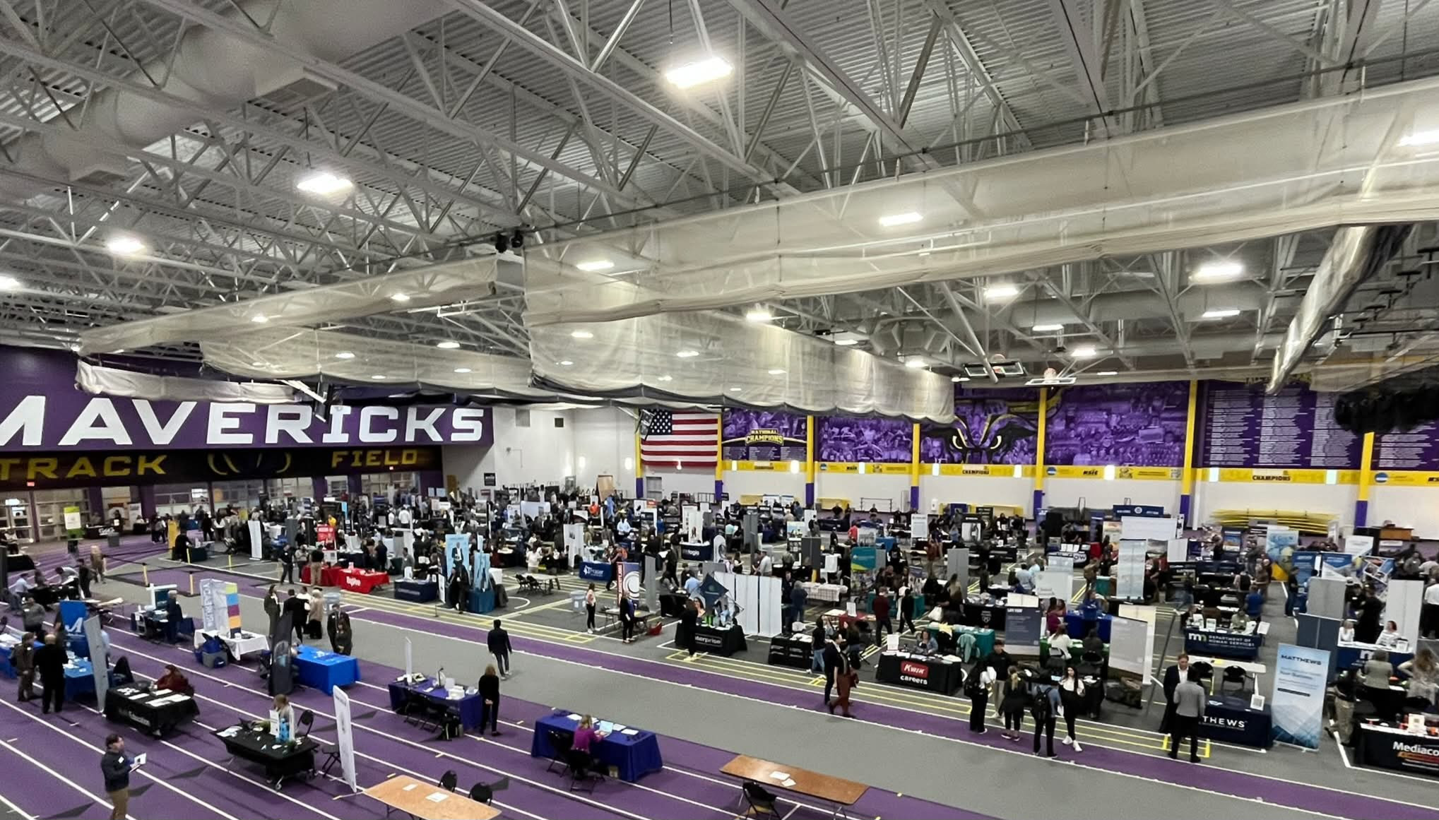 An indoor sports facility hosting a fair, with numerous booths and attendees, under a high ceiling with exposed beams.